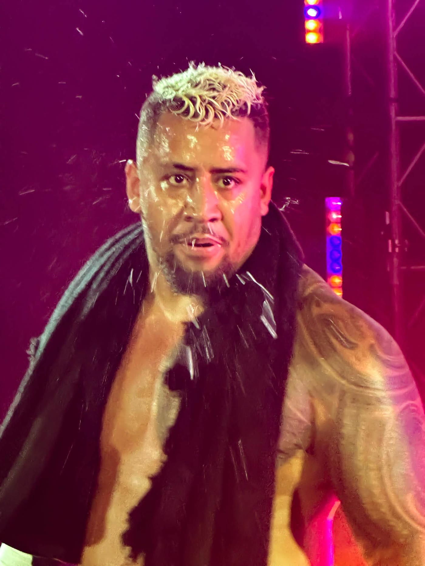 Bleached-blond wrestler in a dark fur-trimmed robe staring down the camera in red entrance lighting