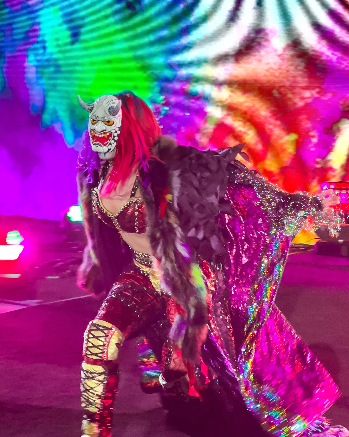 Masked wrestler in a sequinned feathered entrance coat, arms spread against a burst of rainbow smoke