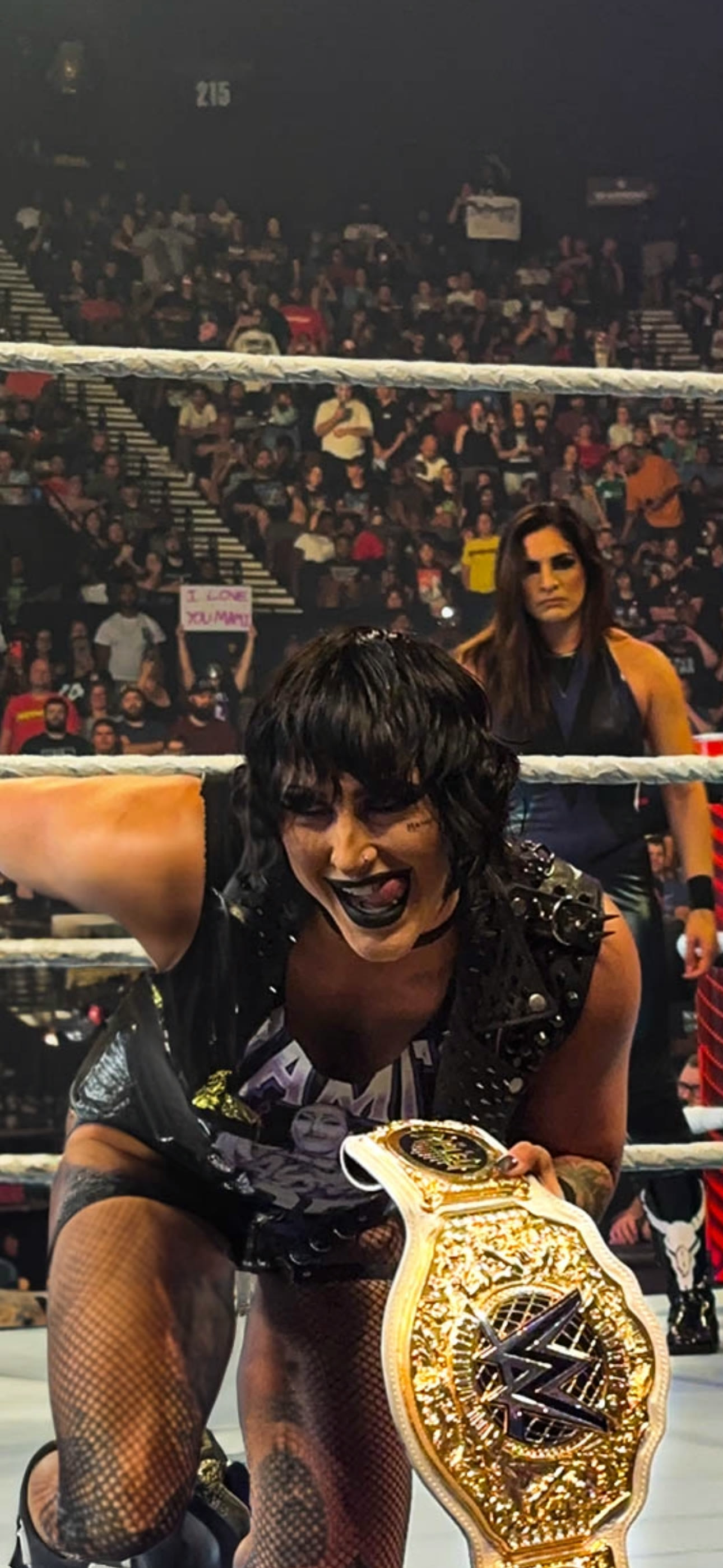 Wrestler kneeling in the ring holding a championship belt out toward a packed crowd